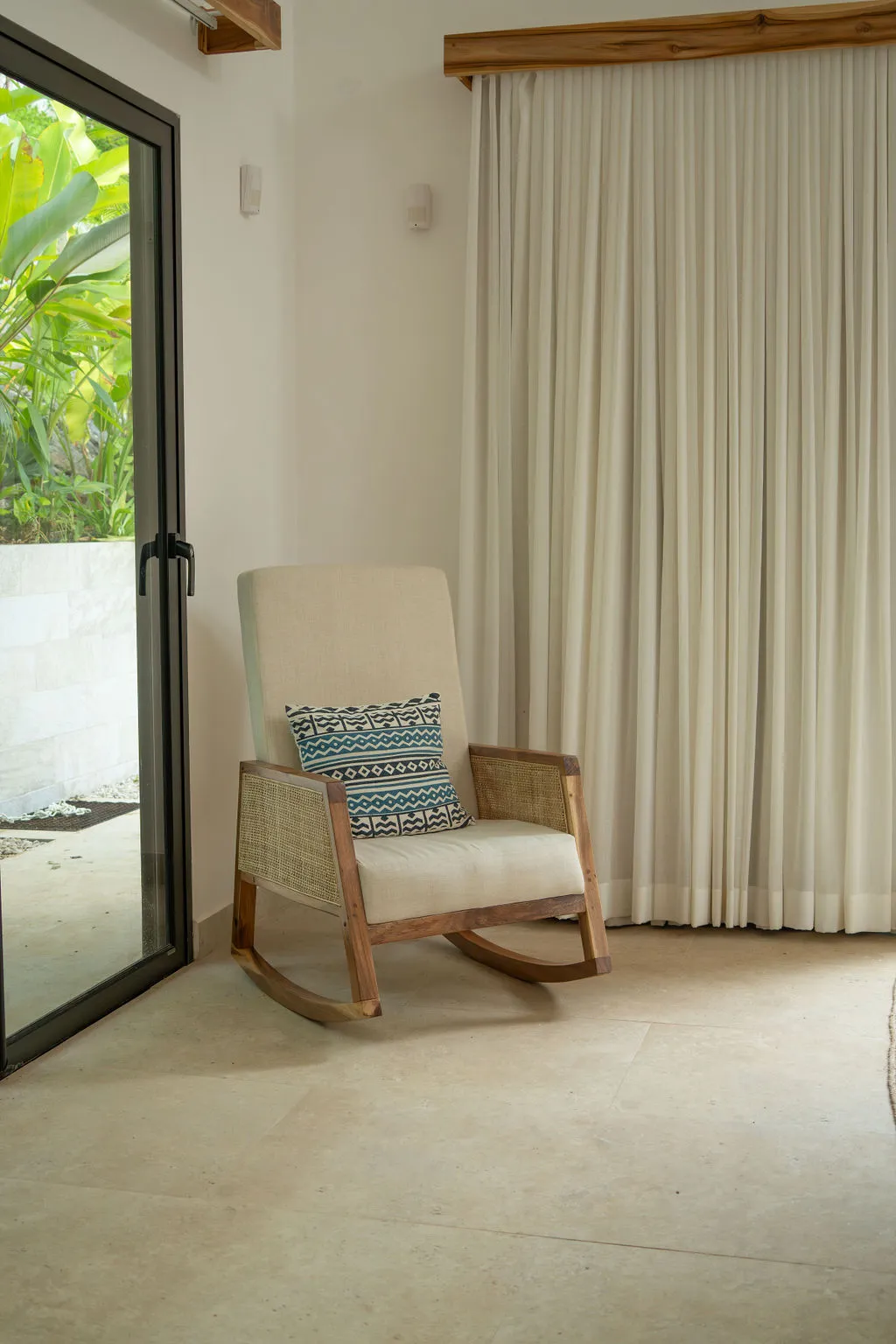 Woven rocking chair beside floor-length curtains and a glass door onto the greenery